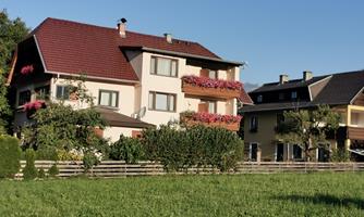 A cozy house with flower boxes on the balconies and surrounding greenery. In the background, more buildings can be seen in a rural setting.