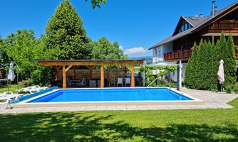 A beautiful garden with a blue swimming pool and a wooden gazebo. Surrounded by green lawn and some trees.