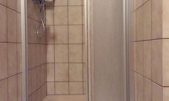 A bathroom with a shower cubicle and tiled walls. On the floor lies a white towel and a plant stands in the corner.