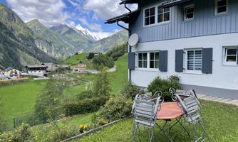 A picturesque house in the mountains with a beautiful garden. In the foreground, there is a table with chairs, and the surroundings are surrounded by impressive mountain landscapes.
