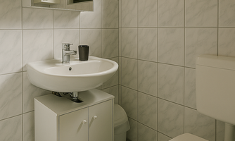 A modern bathroom with white tiles and a small washbasin. There is a window and a simple toilet cabinet.