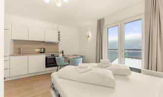 A modern room with a double bed, a kitchenette, and a dining area. Large windows provide a view outside and plenty of daylight.