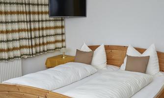 A cozy bedroom with a large wooden bed and white bed covers. A television is mounted on the wall, and the curtains have a nice pattern.