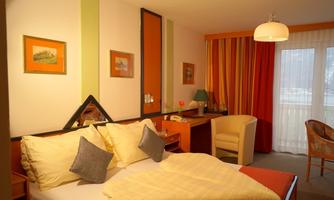 A cozy hotel room with a double bed, desk, and an armchair. The walls are colorfully decorated, and there are large windows with daylight.