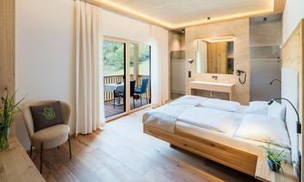 A modern bedroom with wooden decor and large windows. Outside is a balcony with a view of nature.