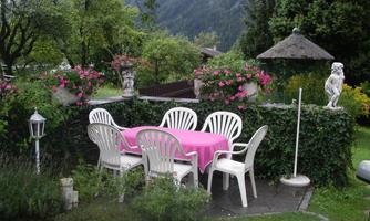 A cozy outdoor area with a pink table and white chairs, surrounded by blooming plants. In the background, trees and a pavilion can be seen.
