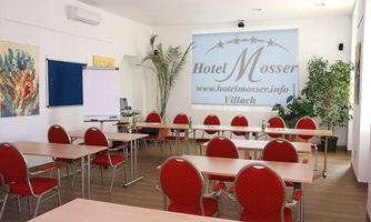 A bright seminar room with red chairs and large tables. There is a projector on the wall and a plant creates a pleasant atmosphere.