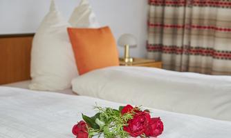 A cozy bedroom with two beds and an orange pillow. On the white bed sheets lies a bouquet of red roses.
