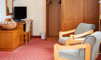 A simple hotel room with red carpet. There is a desk, two armchairs, and a wardrobe.