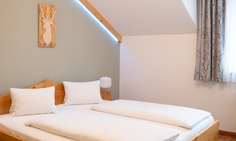 A cozy bedroom with a double bed, white bed linens, and wooden decor. On the wall hangs a depiction of a deer, and the windows are adorned with curtains.