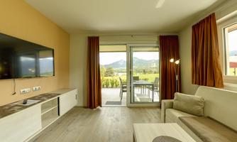A modern living room with a television on the wall and cozy sofa furniture. Large windows offer a view of the landscape with mountains.