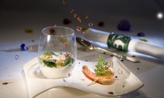 An elegant dish with a glass of appetizer and a shrimp skewer. In the background, decorative elements and a napkin are visible.