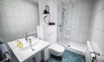 A modern bathroom with white tiles and a glass shower. A sink with toiletries and a towel is also visible.