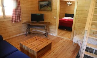 A cozy wooden room with a sofa and a table. In the background, a bedroom with a red bed is visible.