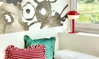 A cozy room with a large window and colorful cushions. In the background, there is an artistic wall design.