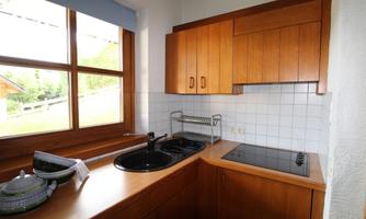 A modern kitchen with wooden furniture and large windows that let in plenty of light. The sink and cooktop are clearly visible on the countertop.