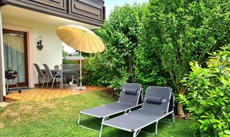 A cozy garden with two loungers and a sunshade. In the background, there is a dining area under a canopy.