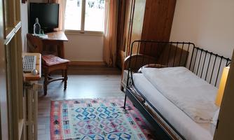 A small room with a single bed, a table, and a television. It has a window with curtains and a colorful carpet on the floor.