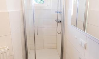 A modern bathroom with a shower and a sink. The walls are light tiled and there is plenty of daylight.