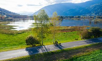 A picturesque landscape with a cyclist on a road. In the background, there is a tranquil lake and gentle hills visible.