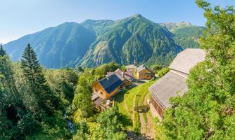 A picturesque mountain village with several wooden houses and lush vegetation. In the background, impressive green mountains rise beneath a clear sky.