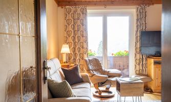 A cozy living room with a comfortable sofa, an armchair, and a small table. Large windows let in plenty of daylight and offer a view outside.