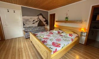 A cozy bedroom with a large wooden bed and colorful flower bedding. A mountain motif can be seen on the wall, and there is a sofa as well as soft lighting.