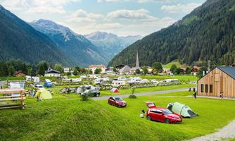 A picturesque campsite in a mountainous landscape. Tents and cars are on a green meadow, surrounded by mountains and a small village.