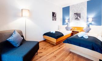 A modern hotel room with two single beds and a cozy sofa. The wall is designed in blue and white, with stylish lighting.