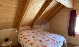 A cozy bedroom with a double bed and wooden paneling. The windows let in plenty of light and there is a small table beside it.