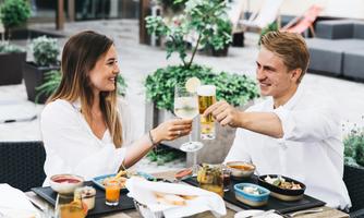 A couple toasts with drinks and enjoys a meal outdoors. The relaxed atmosphere is surrounded by green plants.
