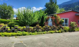 A red house surrounded by lush greenery and colorful plants. In the background, there are mountains and a clear blue sky.