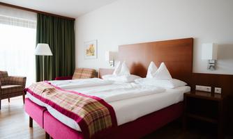 A modern hotel room with a large double bed and cozy decor. The room has windows with curtains and a view of natural light.