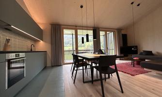 A modern apartment with an open kitchen and a dining area. Large windows provide a view of the nature outside.