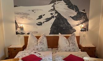 A cozy bedroom with two beds, decorated with cushions in red and a blooming tablecloth. In the background, there is a large mountain motif.