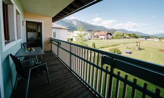 A balcony with a table and a chair that offers a view of green meadows and mountains. The scenery is characterized by a calm, rural atmosphere.