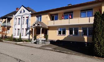 A quiet residential building on a small street. The facade is light yellow with some windows and an awning.