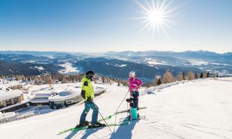 Two skiers are standing on a snow-covered slope with a view of the mountains. Bright sunshine and a clear blue sky create a beautiful winter landscape.