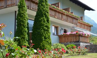 A beautiful building with a balcony and wooden porch. In the foreground, colorful roses are blooming, and there are tall, green cypress trees.