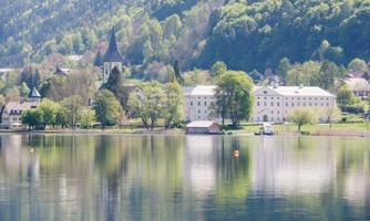 A picturesque landscape with a calm lake and green hills. In the background, a historic building and a church can be seen.
