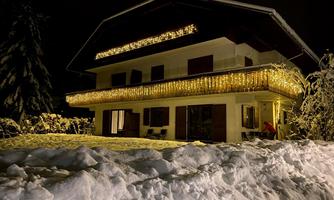 A cozy house at night, decorated with many lights. The snow covers the ground and surrounds the building.
