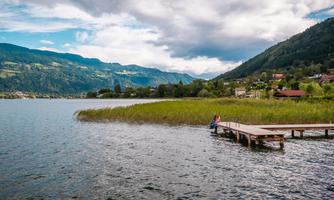 A tranquil lake surrounded by mountains and lush greenery. A person sits on a wooden walkway and enjoys the view.