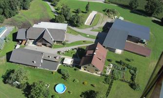 An aerial view of a farm with several buildings and a pool. The surroundings are green and rural.