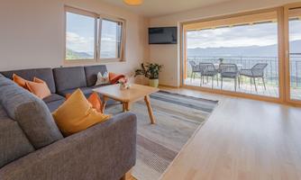 A modern living room with a gray sofa and decorative cushions. The view extends over a terrace with garden furniture and beautiful landscape.