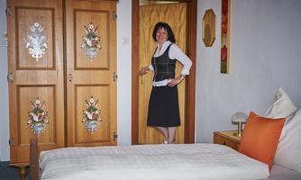 A woman stands in front of a wooden door in a cozy room. The room is furnished with a bed and a wardrobe with floral patterns.