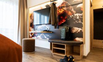 A modern room with a TV on the wall and a padded stool. In the foreground, there is a backpack and a pair of shoes.