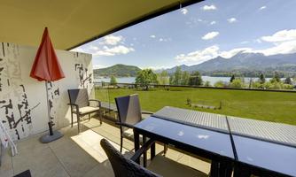 A beautiful terrace with a table and chairs overlooks a picturesque landscape. In the background, there are mountains and a calm lake, under a blue sky with some clouds.