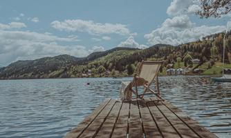 A wooden walkway leads over calm water to a chair. In the background, gentle hills and a clear sky are visible.