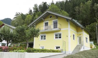 A light yellow house with a balcony and large windows. It is located in a green environment with trees and blooming plants.