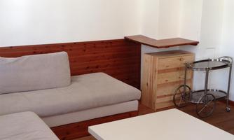 A bright living space with a beige couch and a white table. Pictures are hanging on the wall and there is a wooden table in the corner.